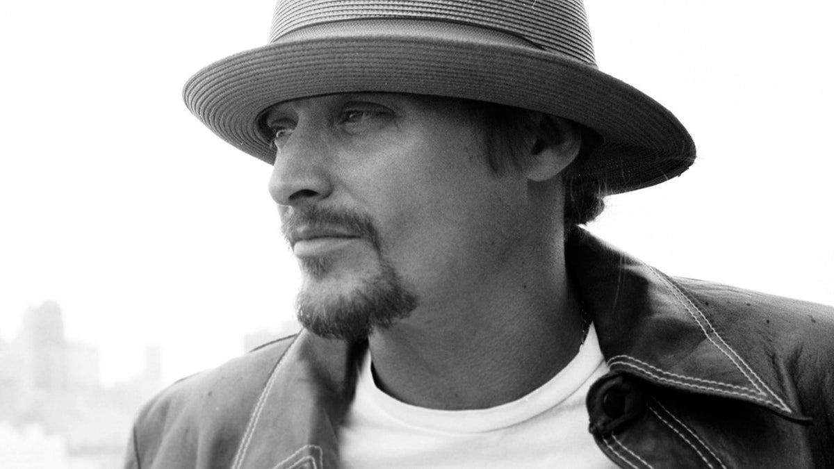 Kid Rock wearing a wide-brimmed hat and dark jacket looks to the left in a black-and-white close-up portrait.