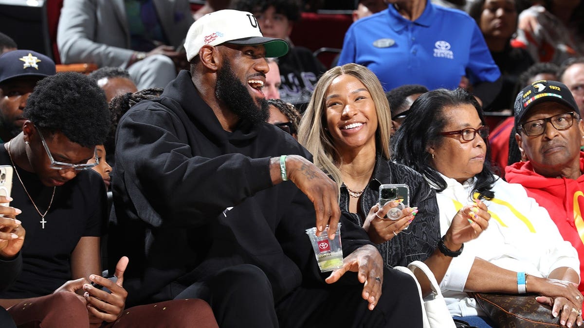 LeBron James, Savannah James and Bryce James sit in the front row at a basketball game.