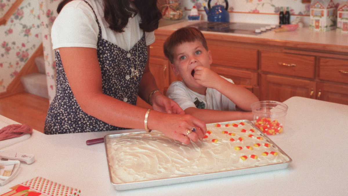 A view of a woman and young boy working together in a kitchen, decorating baked pumpkin bars with candy corn kernels.