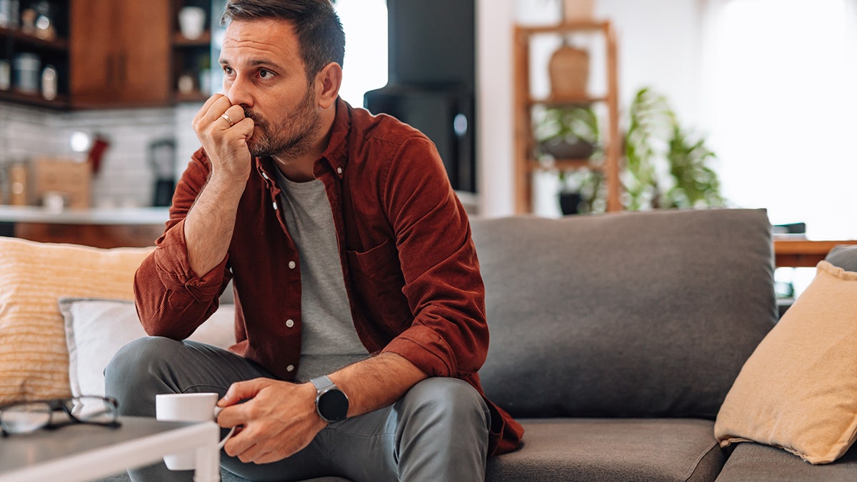 Worried man sitting on the sofa at home, biting fingernails while holding a cup of coffee, deeply lost in thought and grappling with feelings of anxiety and uncertainty