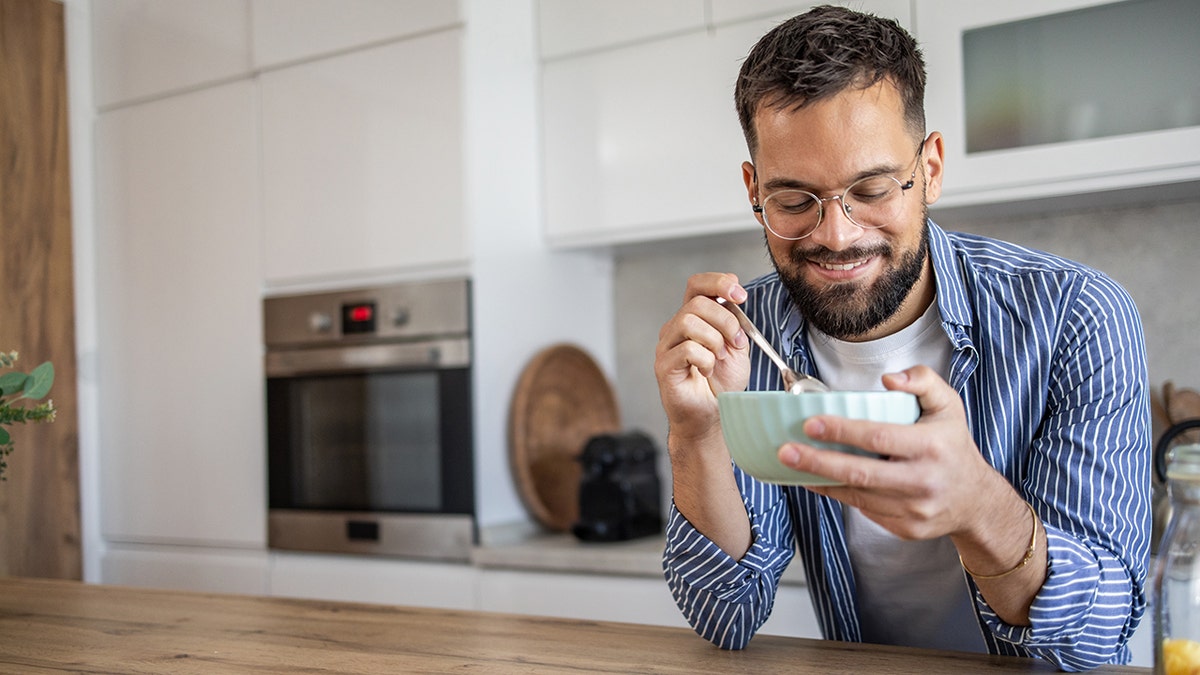 A man wearing glasses eats cereal while leaning on his kitchen island.