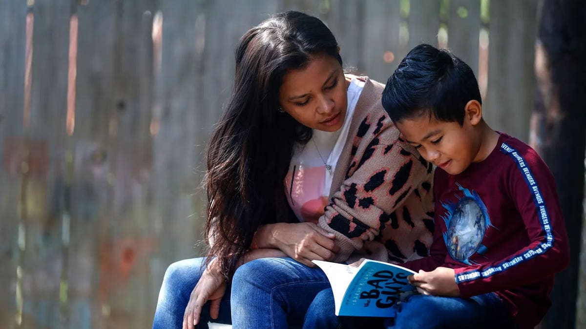 A mother and her son read a book together while sitting outside.