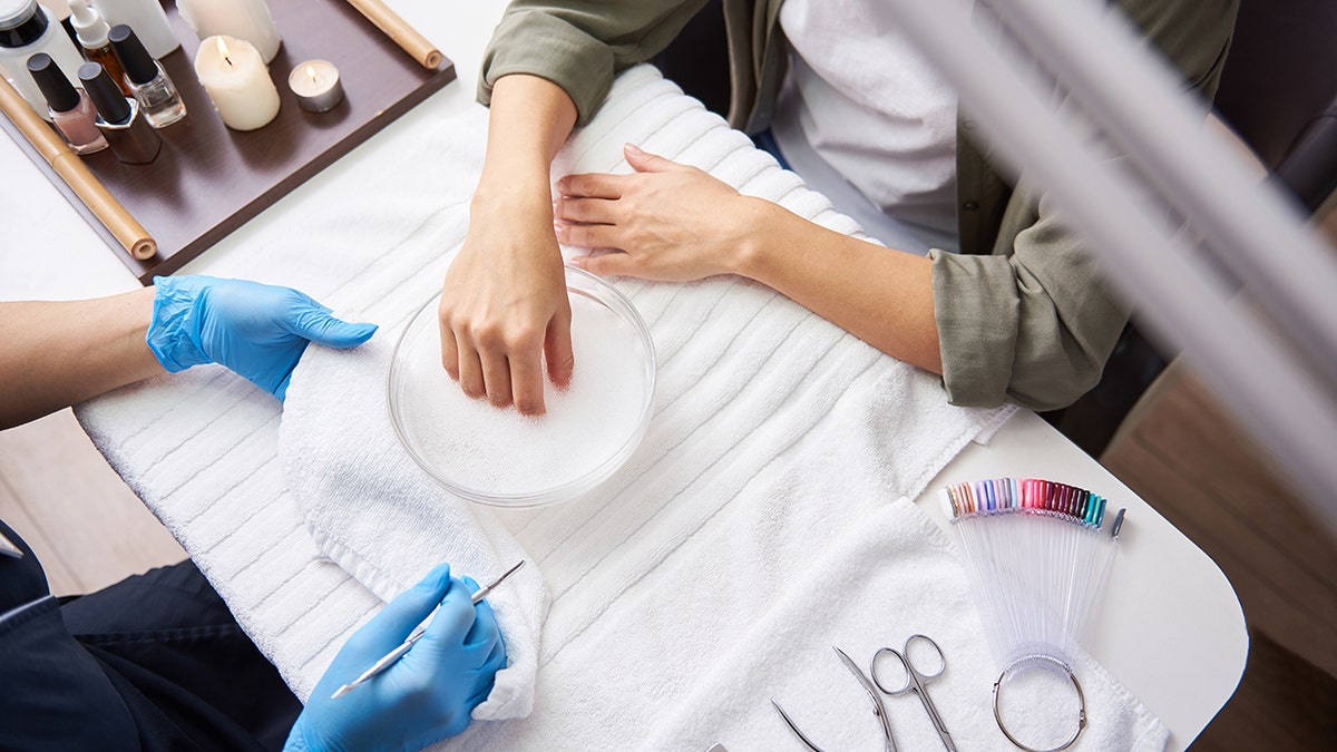 Close up top view picture of young lady soaking arm in nail bath while sitting at the table with manicure tools. Manicurist in sterile gloves holding white soft towel