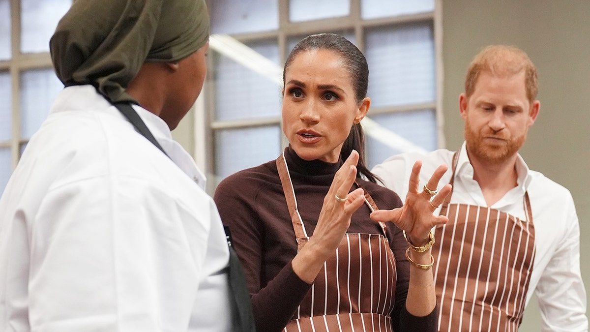 Meghan Markle speaking to someone wearing an apron in a kitchen.