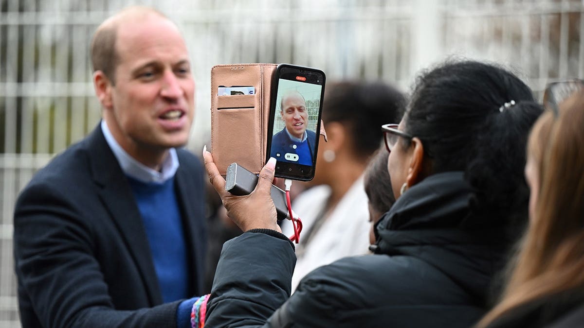 Prince William, Prince of Wales, is filmed on a mobile phone while meeting well-wishers.