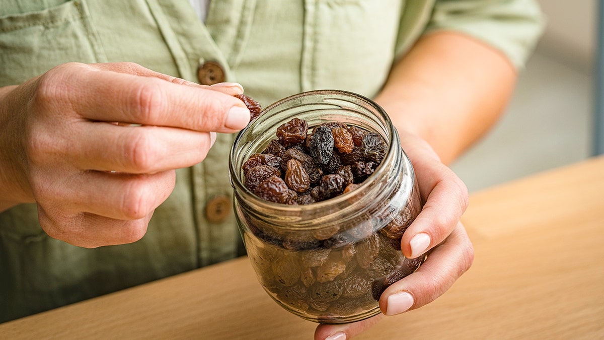 A hand holds a raisin and a jar of raisins in the other.