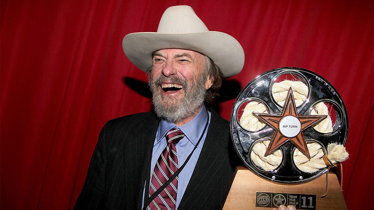 Rip Torn in a white cowboy hat and dark suit laughs beside a star-shaped film award.