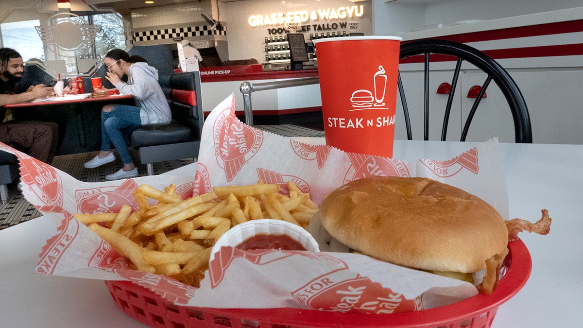 A burger, fries and a drink are shown on a white table at Steak 'n Shake as a man and woman at another table in the top left corner are seen eating.