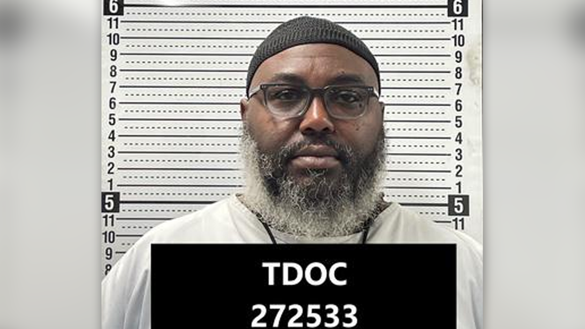 Tadaryl Shipp in a mugshot standing in front of a height board, wearing a white T-shirt, a black beanie, black glasses and a full salt-and-pepper beard