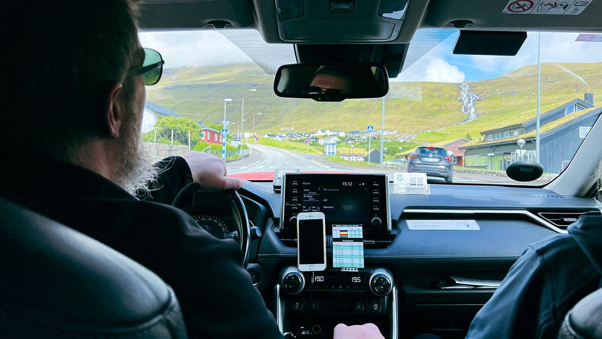 A male taxi driver operates a modern taxi with scenic green mountains visible through the windshield.