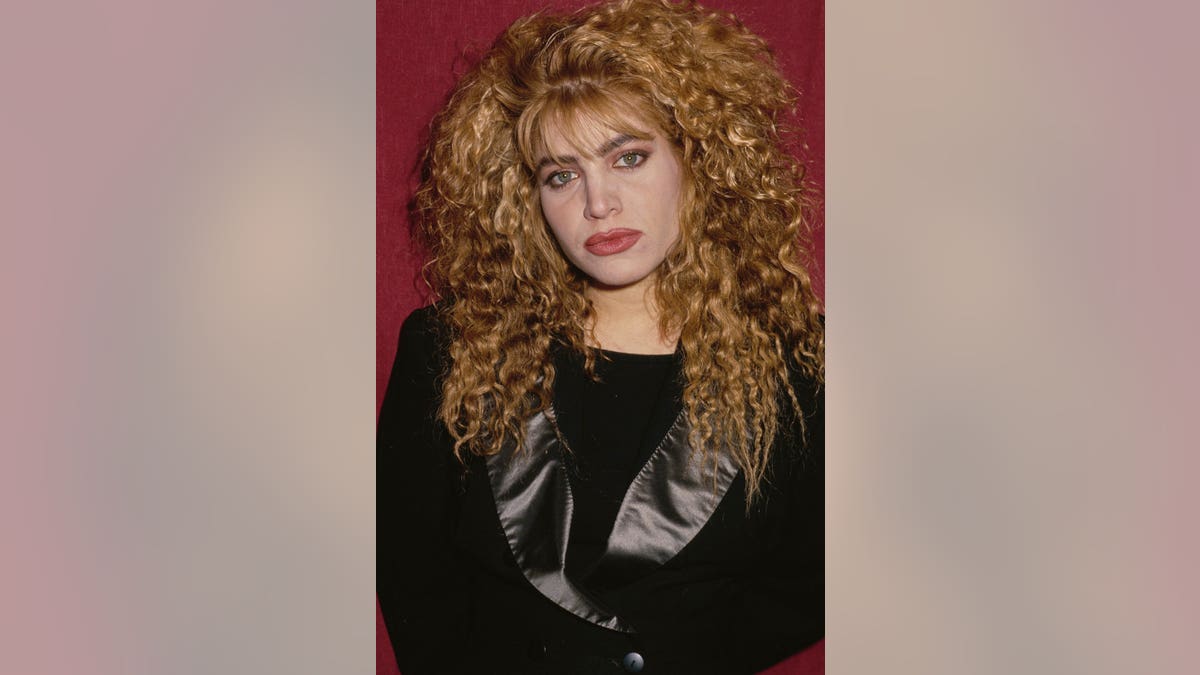 Taylor Dayne wearing a black jacket with silk lapels poses in front of a red backdrop.