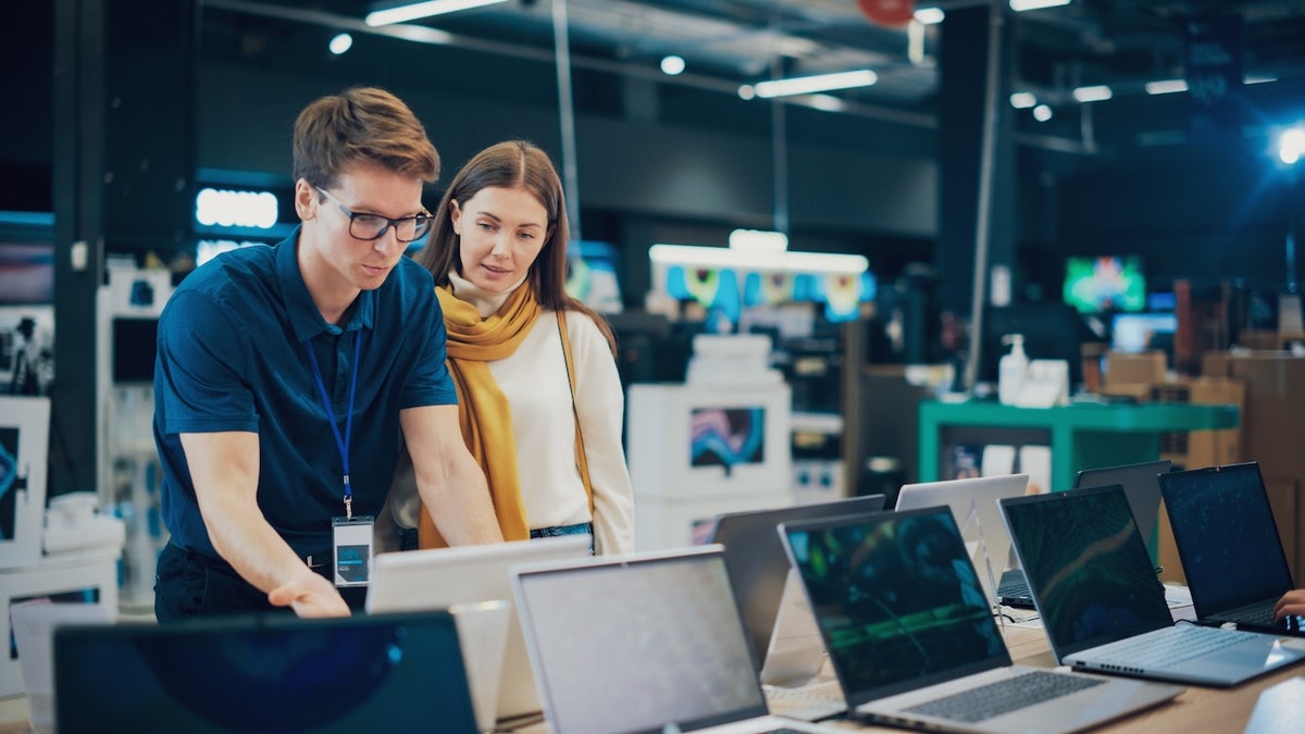 Woman Discusses Laptop Options with Home Electronics Store Sales Associate. Young Shopper Examines Modern Computer Choices with the Help of a Sales Expert in a Showroom.
