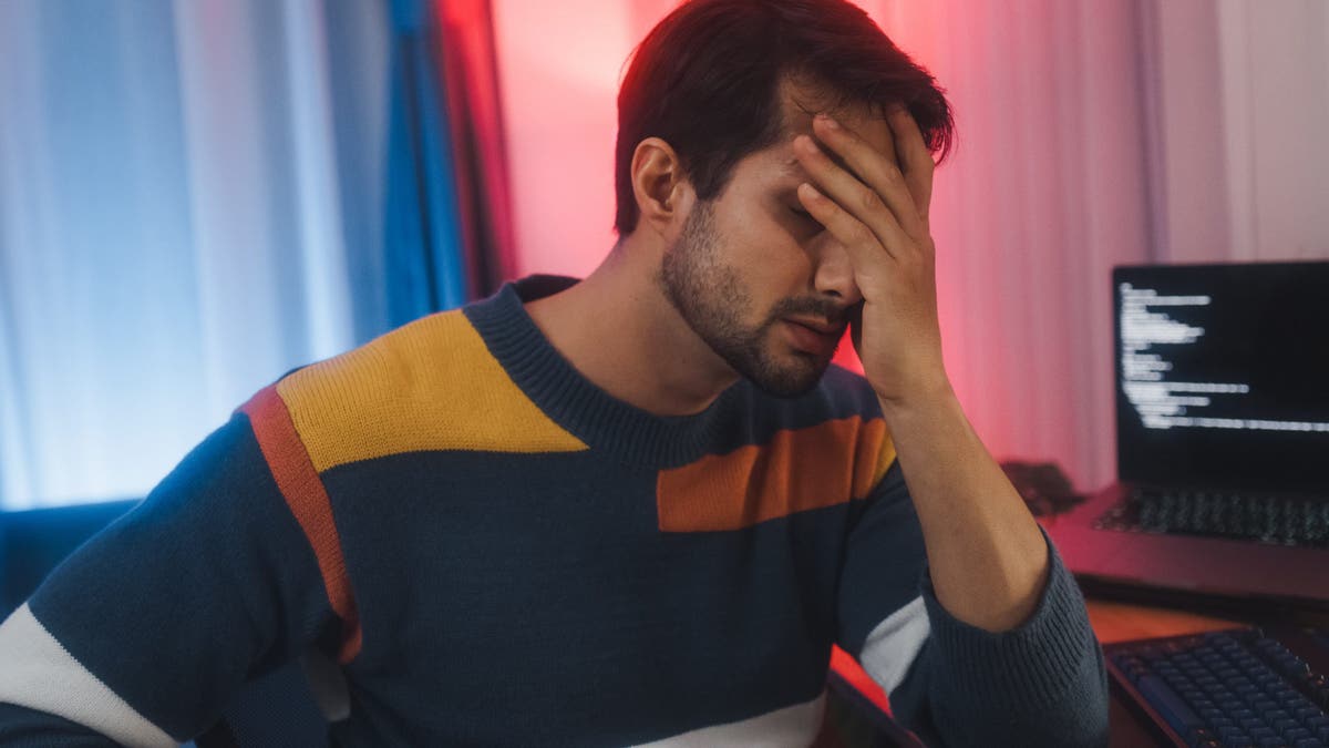 Tired man sits at desk with multiple computer screens in home office at night.