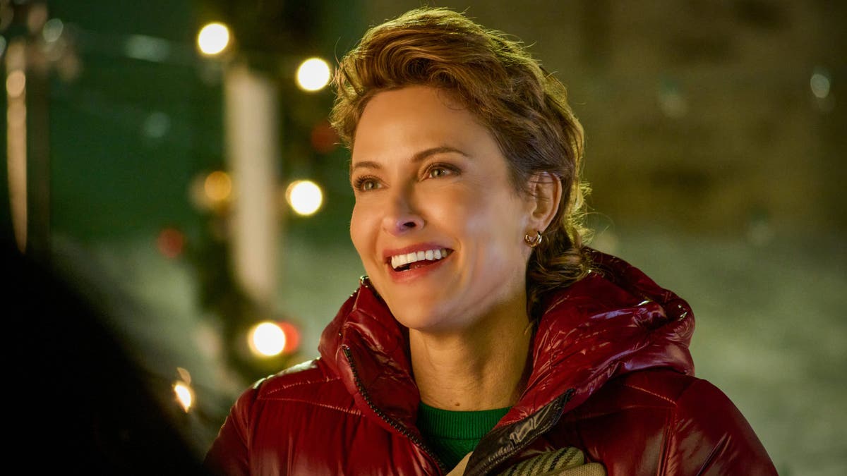 Jill Wagner smiles while wearing a red puffer jacket and patterned gloves in front of softly glowing lights.