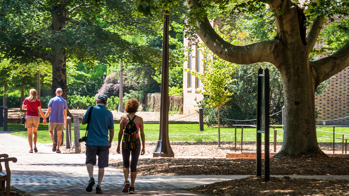 People walk along a shaded brick pathway beside campus buildings at William & Mary in Williamsburg, Virginia