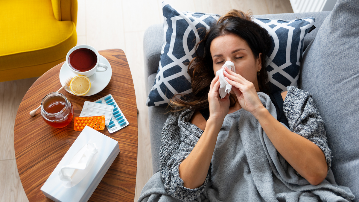 A woman lies on a couch with a tissue in her mouth while a cup of tea sits on a table next to her.
