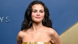 Selena Gomez poses in a strapless gold outfit against a blue backdrop.