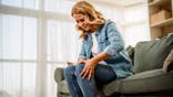 Mature woman sits on a sofa and rubs her knee in pain.