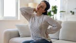 Woman sitting on sofa, holding her neck and lower back in pain.
