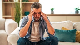 Mature man sitting on the sofa, cradling his head in his hands, grappling with intense headache pain, feeling overwhelmed by stress and exhaustion in a cozy living room setting