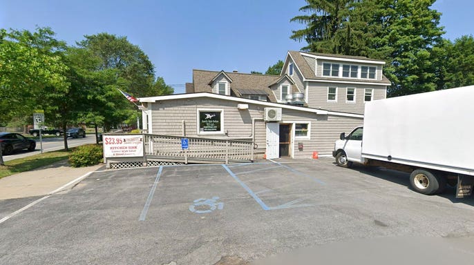 Exterior of a gray-shingled building with a wheelchair ramp, a sign advertising a $23.95 Thursday to-go special, and a white box truck parked beside an empty lot.