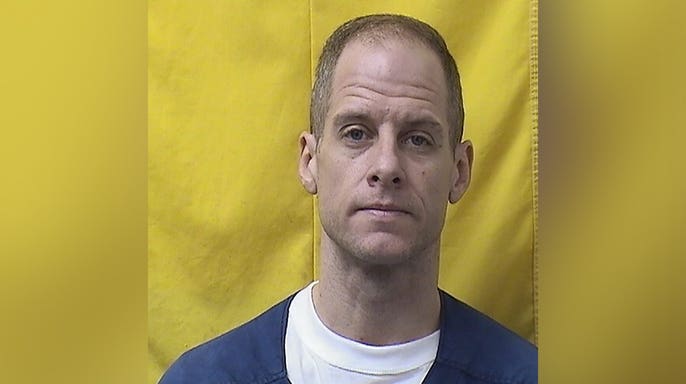 New prison mugshot of convicted wife killer Caleb Flynn wearing a blue prison uniform over a white shirt against a yellow background.