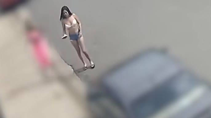 A woman in a light-colored top, denim shorts and sandals faces the camera on a paved street, holding an object in one hand.