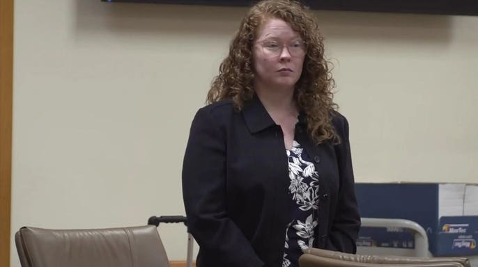Caylen Deller stands in a Florida courtroom wearing glasses, a dark blazer and a floral blouse during proceedings in her boyfriend's murder case.