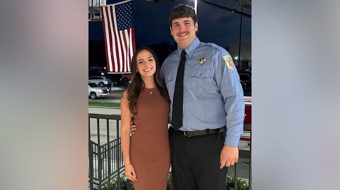 Firefighter Shane Hirstius, wearing a light blue uniform shirt and black tie, stands with his fiancée, Leah Fonte, in a brown dress beneath a large American flag.