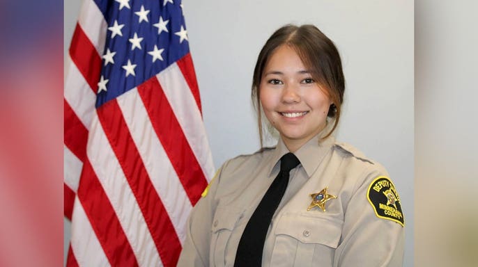 Minnehaha County Sheriff's Deputy Katherine Frank smiles in uniform beside an American flag, wearing a tan shirt, black tie and gold sheriff's badge.
