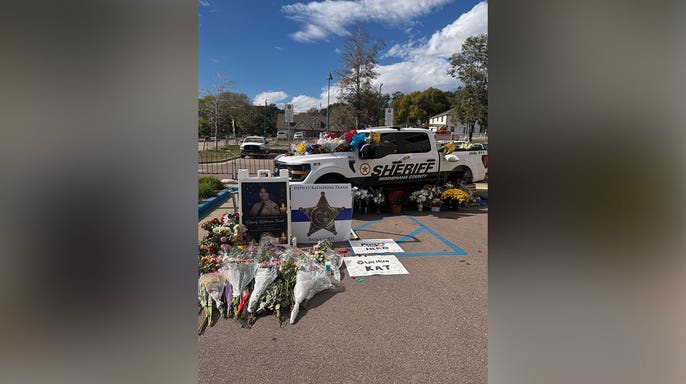 Memorial honoring Deputy Katherine Frank features a white sheriff's pickup truck covered with flowers, framed photographs, bouquets and handwritten tributes outside a law enforcement building.