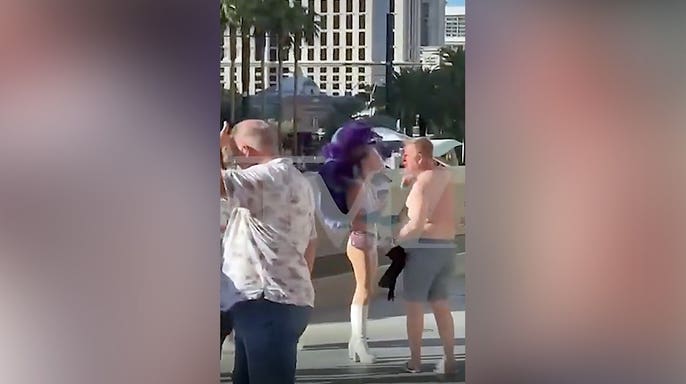 A shirtless man in gray shorts walks away from a woman wearing a white showgirl costume with large purple feathers near an escalator on the Las Vegas Strip.