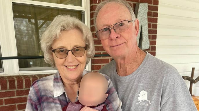 Bobby McGill and his wife, Peggy, smile while holding a young child outside a brick home.