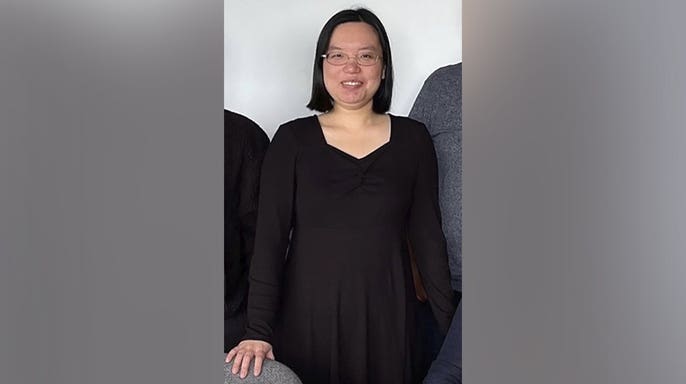 Aili Wang, a University of Wyoming graduate student accused of poisoning a fellow student, stands indoors wearing glasses and a black dress.