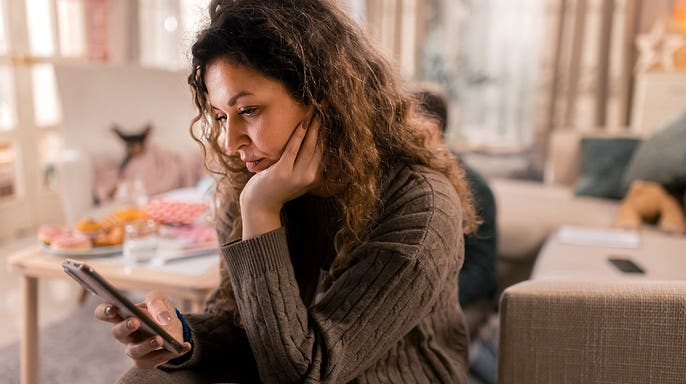 Woman looking worried while checking smartphone at home with family in background.